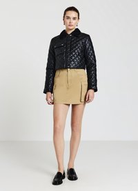 Veste matelassée noire avec un col, poche frontale, associée à une jupe kaki et des mocassins noirs. Texture lisse et design structuré.
