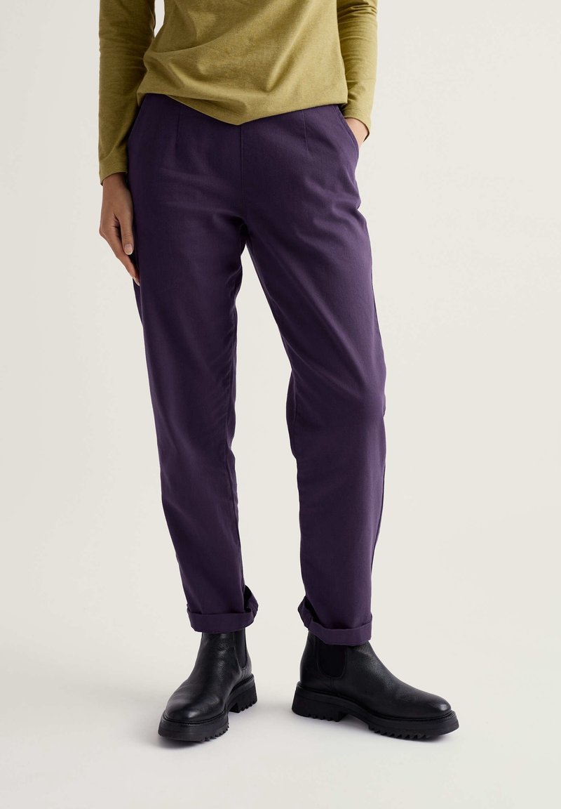 Personne portant un pantalon violet foncé retroussé aux chevilles, des bottines noires et un haut vert à manches longues, une main dans la poche.