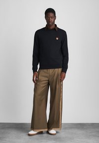 Črna majica s kapuco z majhnim oranžnim vezenim logotipom, v kombinaciji s bež hlačami širokih nogavic in belimi športnimi copati. Gladka tekstura tkanine.