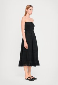 Vestido preto sem alças com corpete texturizado e bainha com camadas de volantes. Usado com mules pretos planos. Silhueta simples e elegante.