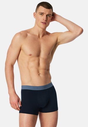 Homme torse nu aux cheveux courts portant un boxer sombre avec une ceinture bleue, se touchant la tête, sur un fond blanc uni.