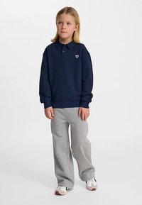 Marinblå pikétröja med krage och liten logotyp, kombinerad med ljusgråa vidbeniga mjukisbyxor och vita sneakers med gröna detaljer.