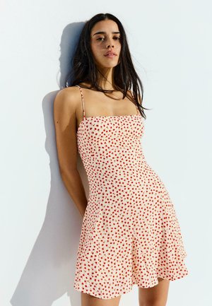 Mladá žena s dlhými tmavými vlasmi stojí opretá o bielu stenu, oblečená v bielych šatách s tenkými ramienkami a drobným červeným kvetinovým vzorom.