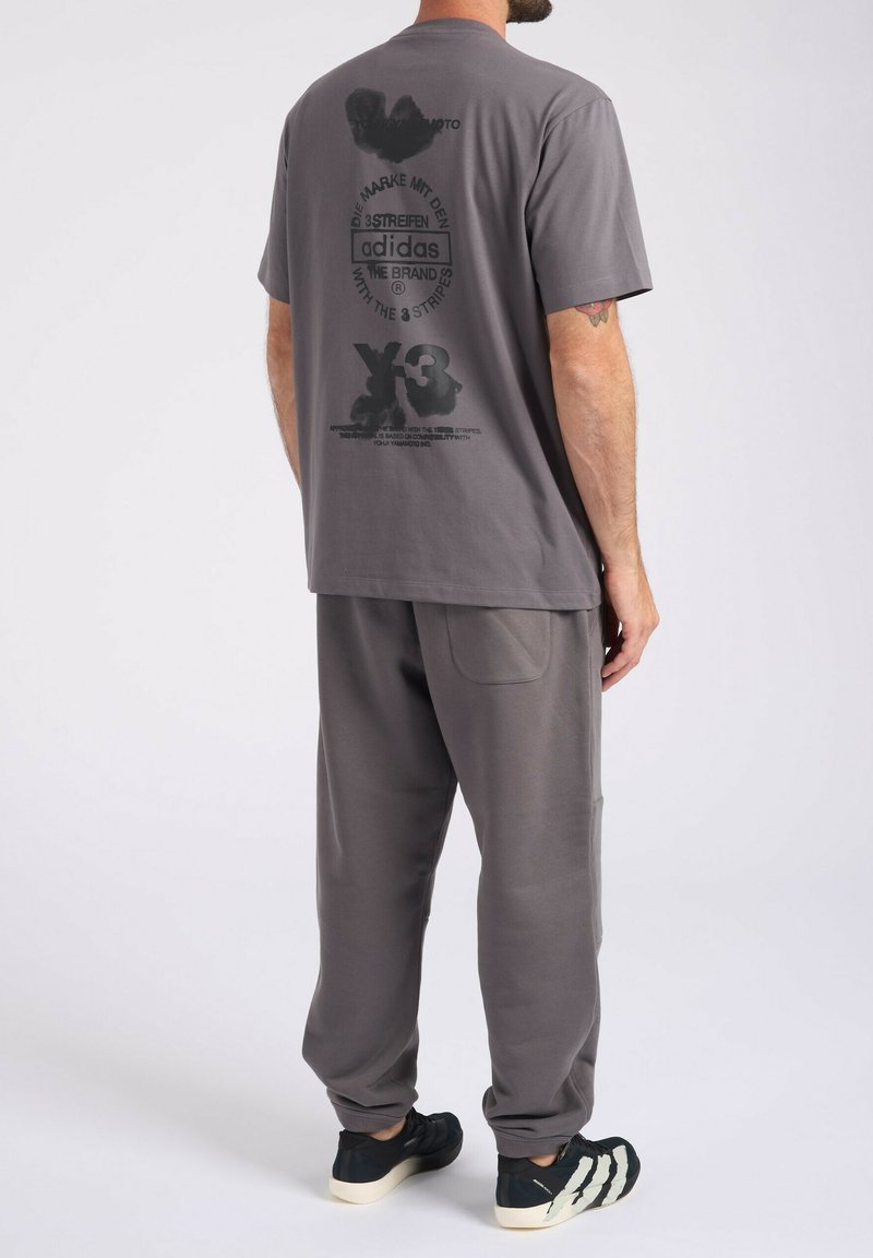 Uomo che indossa una t-shirt grigia Adidas Y-3 con testo nero e simboli sul retro, pantaloni grigi abbinati e sneakers nere con suole bianche.