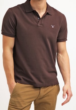 Mand iført en mørkebrun kortærmet polo med et lille logo og beige bukser, står med den ene hånd i lommen.