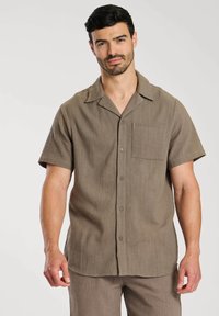Chemise marron à manches courtes en tissu texturé, dot�ée d'un col, de boutons sur le devant et d'une poche poitrine. Ourlet arrondi.