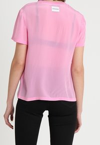 Różowa, przezroczysta bluzka z krótkim rękawem, o prostym kroju i okrągłym dekolcie. Posiada subtelną teksturę oraz metkę z tyłu. Połączona z czarnymi spodniami.