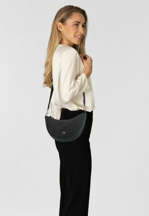Black leather shoulder bag with a curved design, silver hardware, and a smooth texture. Model wearing a cream blouse and black pants.