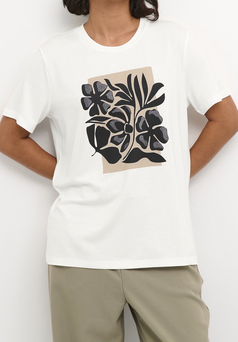 Camiseta blanca con un gran estampado floral negro sobre un fondo beige. Mangas cortas, cuello redondo y textura de tela suave.