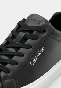 Svart skinnsneaker med en glatt finish, svarta snörningar, en vit sula och "Calvin Klein"-logotyp i vitt längs sidan.