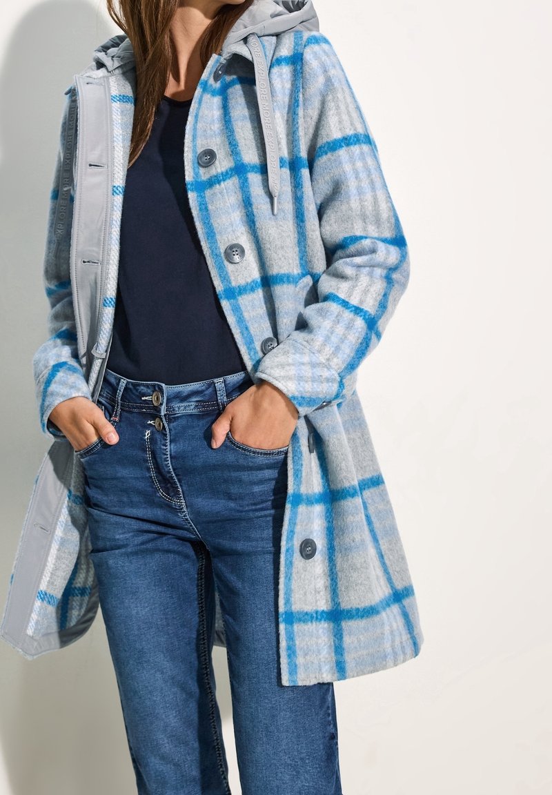 Kariertem Mantel in Grau und Blau mit Kapuze, der eine wollähnliche Textur aufweist, graue Knöpfe und eine entspannte Passform hat. Kombiniert mit blauen Jeans.