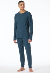 Hombre de pie descalzo, vistiendo un conjunto de camiseta de manga larga y pantalones azules a juego, mirando ligeramente hacia un lado sobre un fondo blanco.
