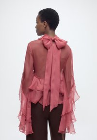 Γυναίκα με την π λάτη γυρισμένη, φοράει διάφανη ροζ μπλούζα με μεγάλο φιόγκο και στρωτά βολάν στα μανίκια, σε συνδυασμό με σκούρο παντελόνι πάνω σε λευκό φόντο.