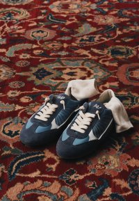 Paire de baskets bleues et blanches avec des lacets crème et des chaussettes blanches à l’intérieur, posées sur un tapis rouge à motif.