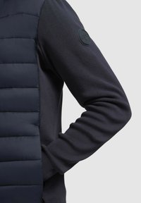 Marineblå sweater med en quiltet puffer-front, ribbede strikærmer og et cirkulært logo-patch på venstre albue.