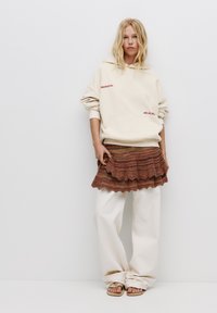 Felpa con cappuccio beige con testo rosso, indossata sopra una gonna a balze marrone e arancione a fantasia, abbinata a pantaloni bianchi larghi e sandali semplici.