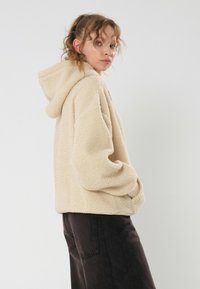 Felpa beige con superficie morbida e texturizzata, tasche a canguro e un ampio cappuccio, abbinata a pantaloni scuri a gamba larga.