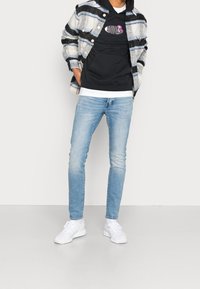 Pohodový outfit obsahující vzorovanou fleecovou bundu v modré a bílé barvě, černou mikinu s logem, světle modré džíny a bílé tenisky.