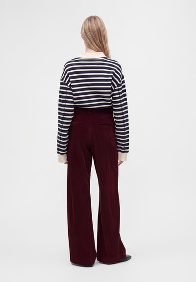 Femme aux longs cheveux blonds portant un haut rayé noir et blanc à manches longues et un pantalon large taille haute rouge foncé, debout de dos.