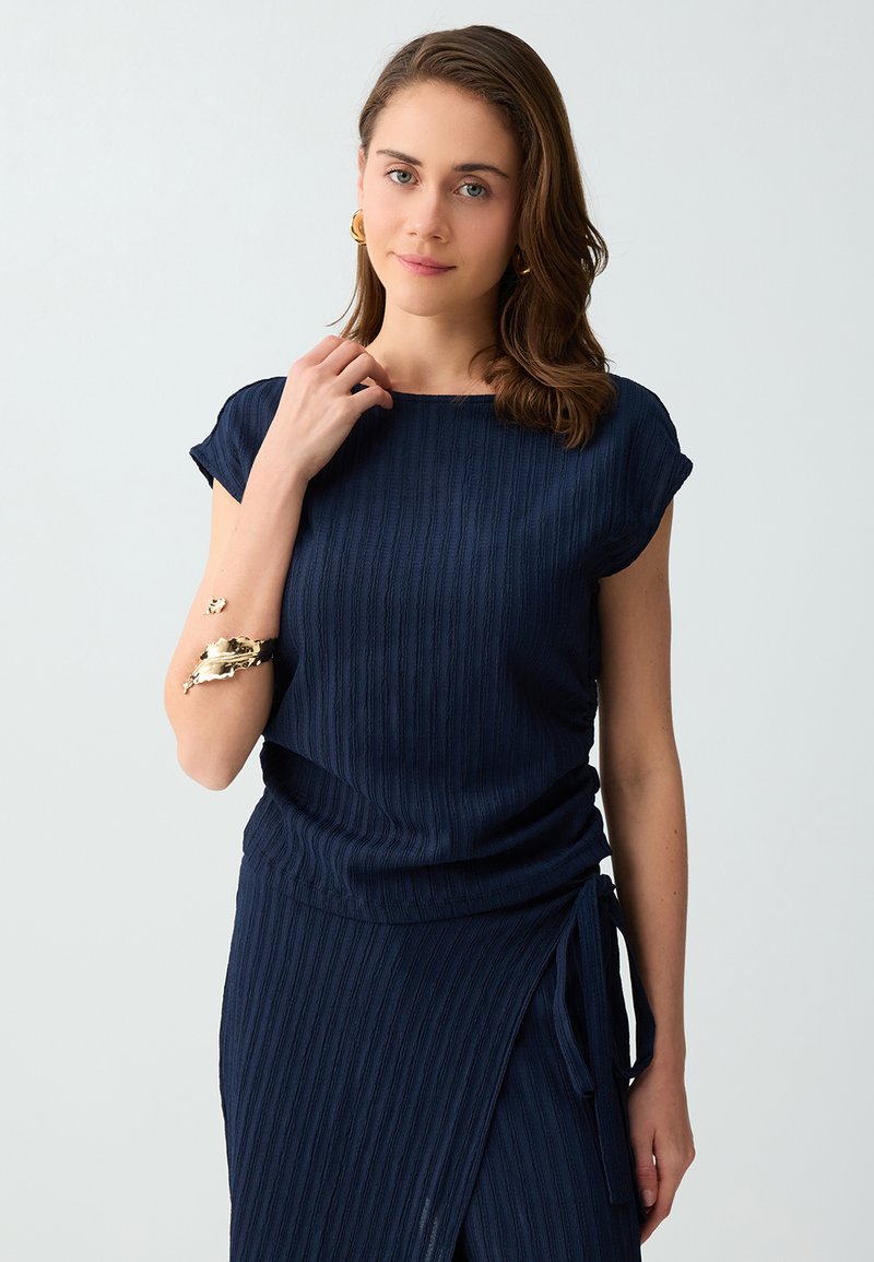 Femme aux cheveux châtain moyen porte une robe côtelée bleu marine à manches courtes et des bijoux en or, debout devant un fond gris clair.