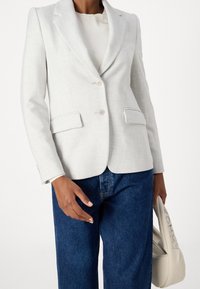 Ljusgrå blazer i ett strukturerat tyg, med notch-lapel, två knappar och framfickor, kombinerad med mörkblå jeans.