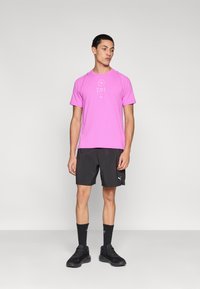 Ανοιχτό ροζ αθλητική μπλούζα με κοντά μανίκια και λογότυπο, συνδυασμένη με μαύρο σορτς και μαύρα αθλητικά παπούτσια, σε ουδέτερο φόντο.