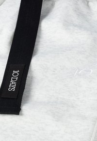 Hellgrauer Stoff mit einem subtilen Muster, ausgestattet mit einem schwarzen Riemen und einem Etikett mit der Aufschrift "10DAYS" in weißer Stickerei. Sauberes, modernes Design.
