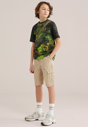 Dítě nosící zelené tričko s grafikou krokodýla, béžové cargo šortky, bílé ponožky s pruhy, tenisky a crossbody tašku, stojící s rukama v kapsách.