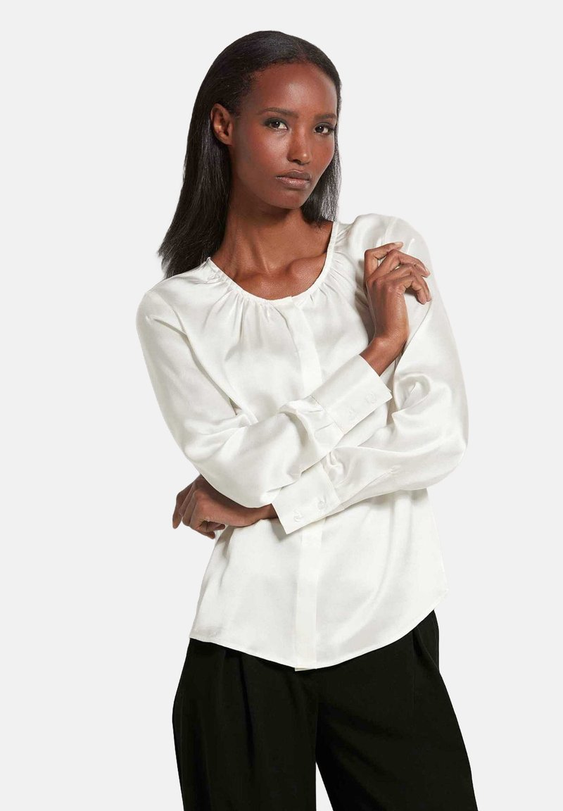 Witte blouse met lange mouwen, een geraffineerde halslijn en knoopdetails op de manchetten, gecombineerd met zwarte broek. Soepele, satijnachtige textuur.