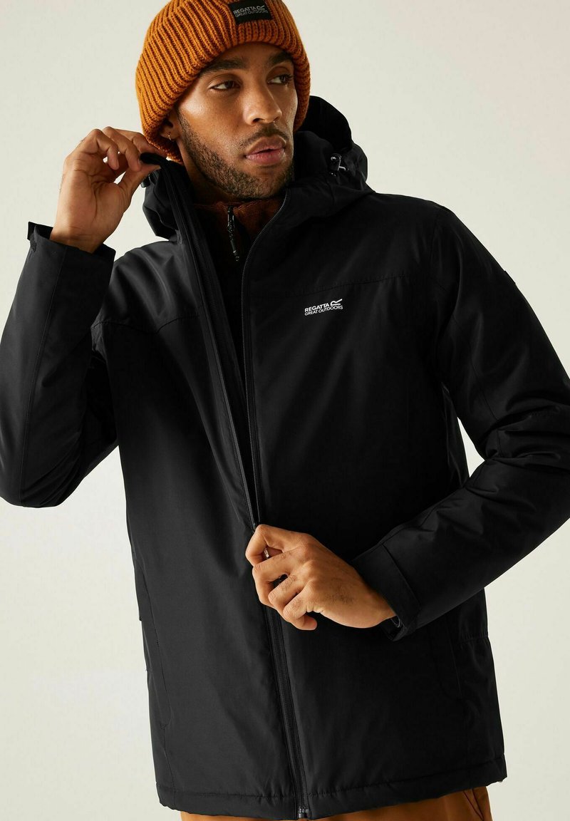 Homme portant un bonnet orange en train de fermer une veste noire Regatta Great Outdoors avec capuche, sur fond uni.