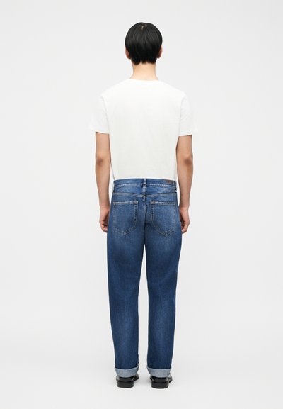 Άτομο με κοντά μαύρα μαλλιά, φορώντας λευκό κοντομάνικο και γαλάζια τζιν με γυρισμένα μπατζάκια, στέκεται πλάτη σε ένα απλό ανοιχτό γκρι φόντο.