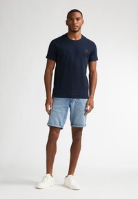 Camiseta de manga corta texturizada en azul marino con un pequeño logotipo en el pecho, combinada con pantalones cortos de mezclilla azul claro remangados y zapatillas blancas.