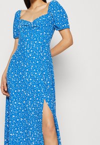 Niebieska kwiatowa sukienka maxi z krótkimi bufkowymi rękawami, z kwadratowym dekoltem i bocznym rozcięciem, wykonana z lekkiego materiału o gładkiej fakturze.