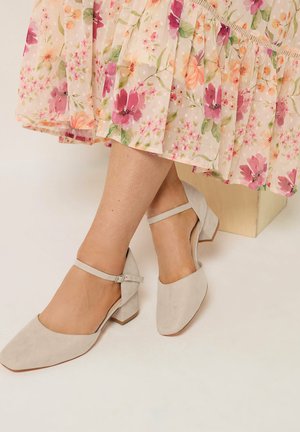 Vrouw die beige suède Mary Jane schoenen met lage hak draagt, gecombineerd met een licht gebloemde midi-rok, zittend op een licht houten blok.