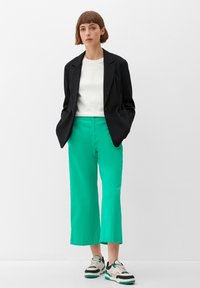 Schwarzes Blazer, weißes T-Shirt und leuchtend grüne, weit geschnittene Hose. Die Sneakers haben grüne, schwarze und cremefarbene Akzente mit einem gestaffelten Design.