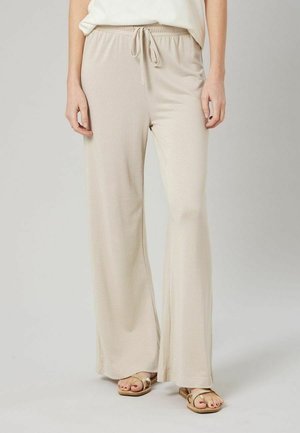 Femme portant un pantalon large beige clair à taille coulissante et des sandales plates dorées, debout devant un fond clair uni.