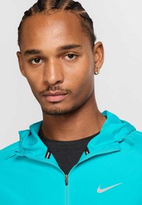 Nike blauwe jas met ritssluiting, capuchon en een zwarte binnenlaag. Voorzien van een zilveren logo en een zachte stoftextuur.