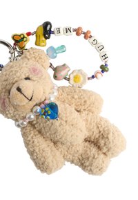 Miękki, beżowy breloczek w kształcie misia, z fakturowanym futrem, ozdobiony perłowym naszyjnikiem i kolorowymi brelokami z napisem "PRZYTUL MNIE" na koralikowym pierścieniu.