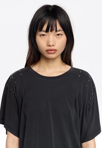 Camiseta negra oversized de tela suave con mangas cortas, que presenta adornos de tachuelas plateadas en los hombros y un cuello redondo.