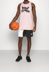 Hombre sosteniendo un balón de baloncesto, vistiendo una camiseta sin mangas rosa con el texto "NO TIRES LA TOALLA", pantalones cortos en blanco y negro y zapatillas negras con acentos neón.