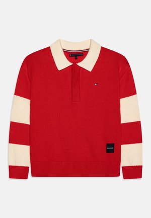 Pull en maille rouge à manches longues avec col crème et larges rayures crème sur les manches, comportant un petit logo brodé sur la poitrine et une étiquette près de l'ourlet.