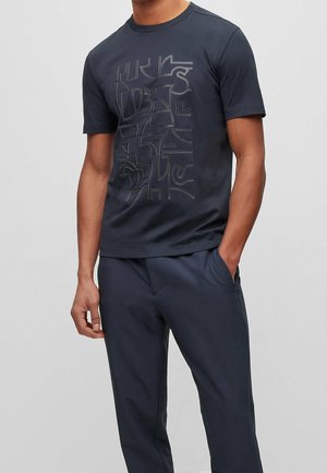 Mężczyzna ubrany w granatowy t-shirt z abstrakcyjnym tłoczonym wzorem i pasujące granatowe spodnie, ręce luźno wzdłuż ciała.