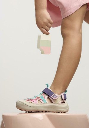 Gamba di bambino con sandalo colorato a strappo che calpesta una superficie rosa, tenendo un giocattolo di legno a forma di gelato con strisce pastello.