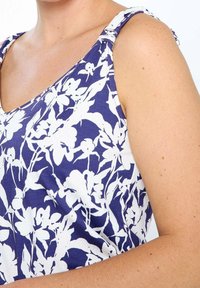 Gros plan sur une personne portant un haut sans manches bleu avec des motifs floraux blancs et des bretelles nouées sur les épaules.