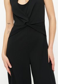 Μαύρη ολόσωμη φόρμα χωρίς μανίκια με λεπτομέρεια σγουρού κόμπου στη μέση, φορεμένη από άτομο με τατουάζ και στα δύο χέρια.