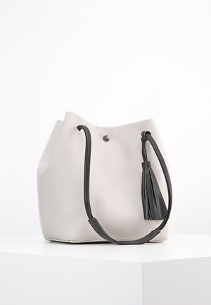 Sac seau blanc avec bandoulière noire réglable et pompon noir assorti, posé sur un socle blanc sur un fond uni.
