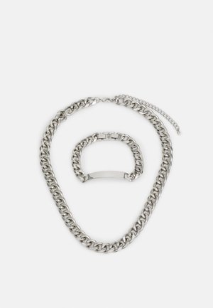 Zilveren metalen chunky ketting en armband set, met in elkaar grijpende schakels en een gladde, gepolijste afwerking. Sluiting detail zichtbaar.