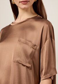 Blusa in satin marrone con una vestibilità ampia, scollo rotondo e maniche corte. Presenta una tasca applicata sul lato sinistro e una texture liscia.