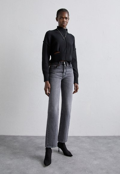 Black cropped sweater with long sleeves and cut-out detail, paired with high-waisted grey jeans and black heeled ankle boots.