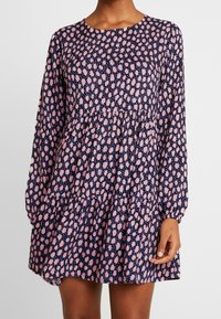 Mörkblå klänning med långa ärmar, rund halsringning och en volangkant i flera lager. Har ett rosa abstrakt mönster genom hela. Slät tygtextur.